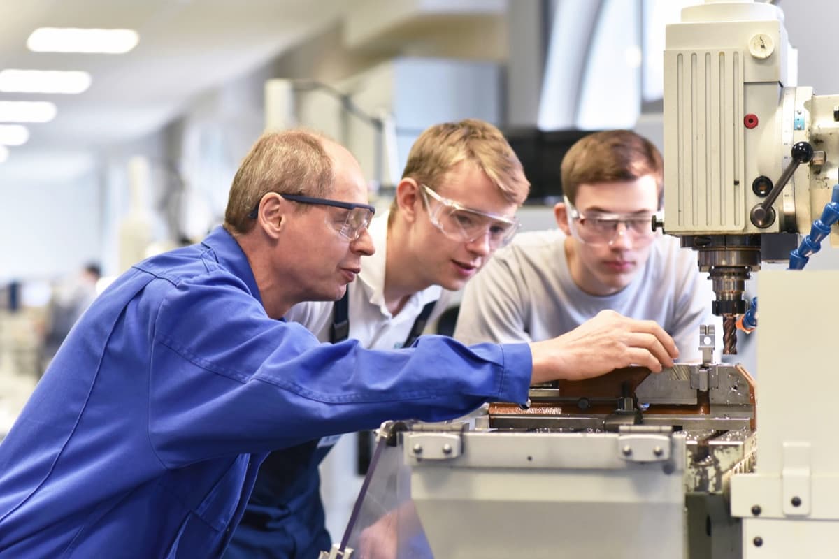 Instructor guiding two students in safety glasses at a milling machine.