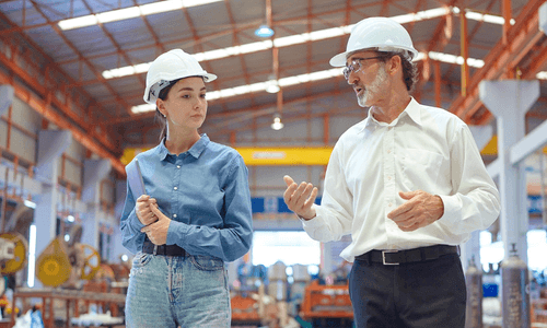 Two colleagues in hard hats talking together on a manufacturing floor.